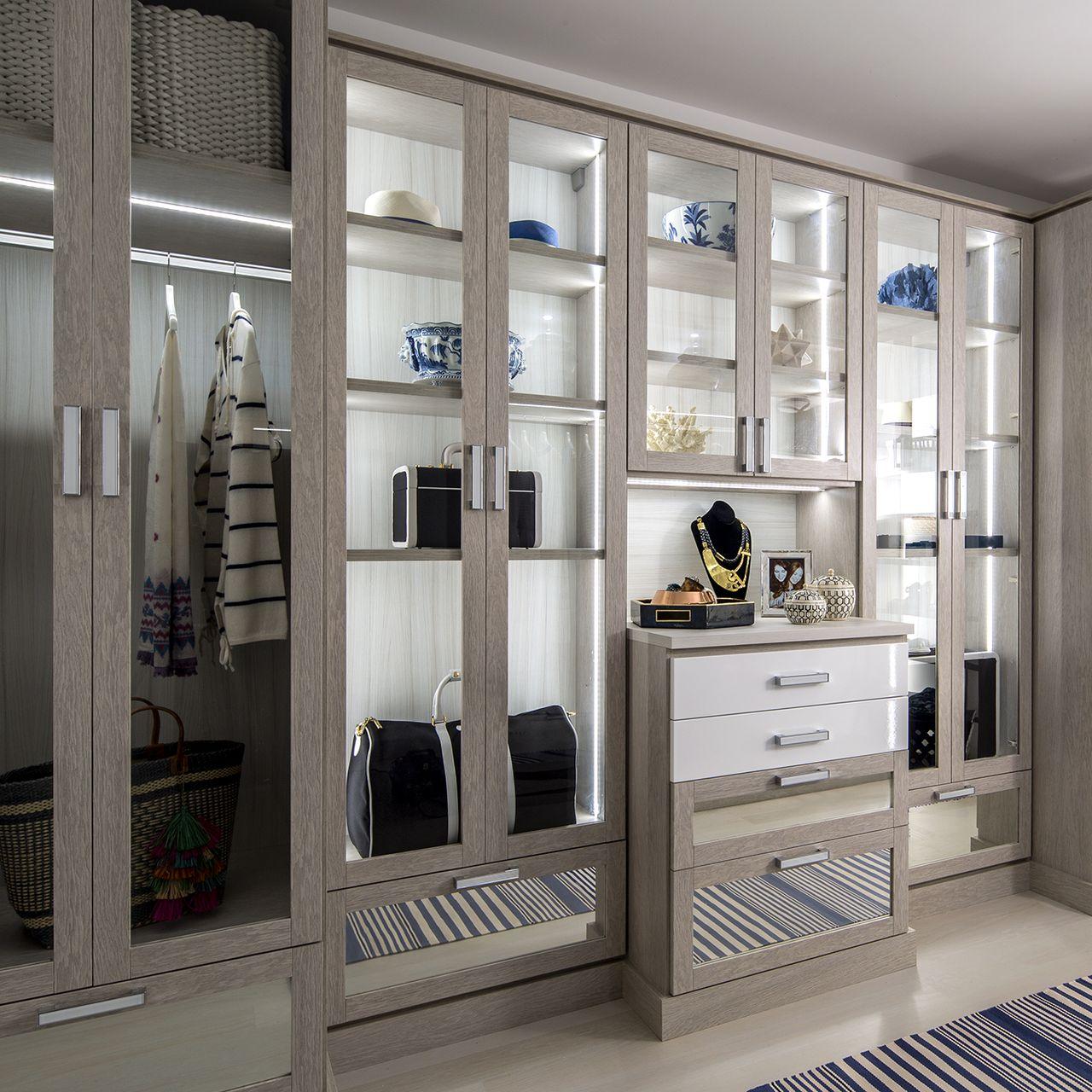 Armario diseñado con gabinetes con puertas de cristal e iluminación por California Closets