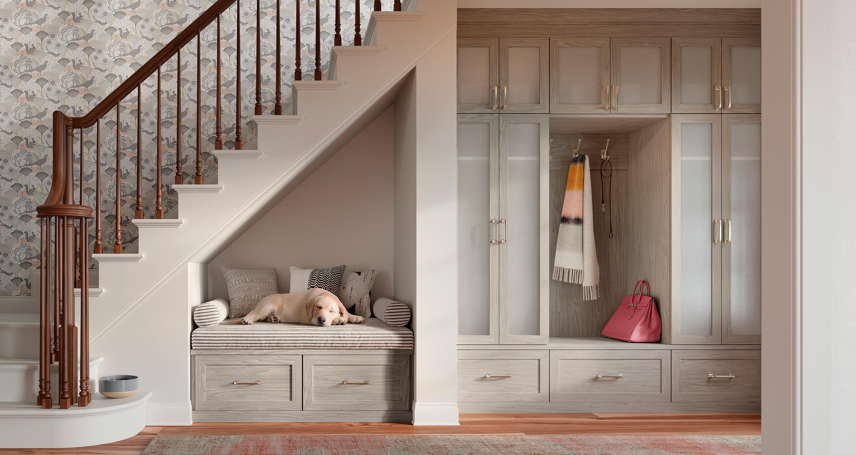 A cozy under stair storage setup by California Closets featuring a cushioned bench with a sleeping dog, built-in cabinets, and hanging space for coats and accessories. | California Closets
