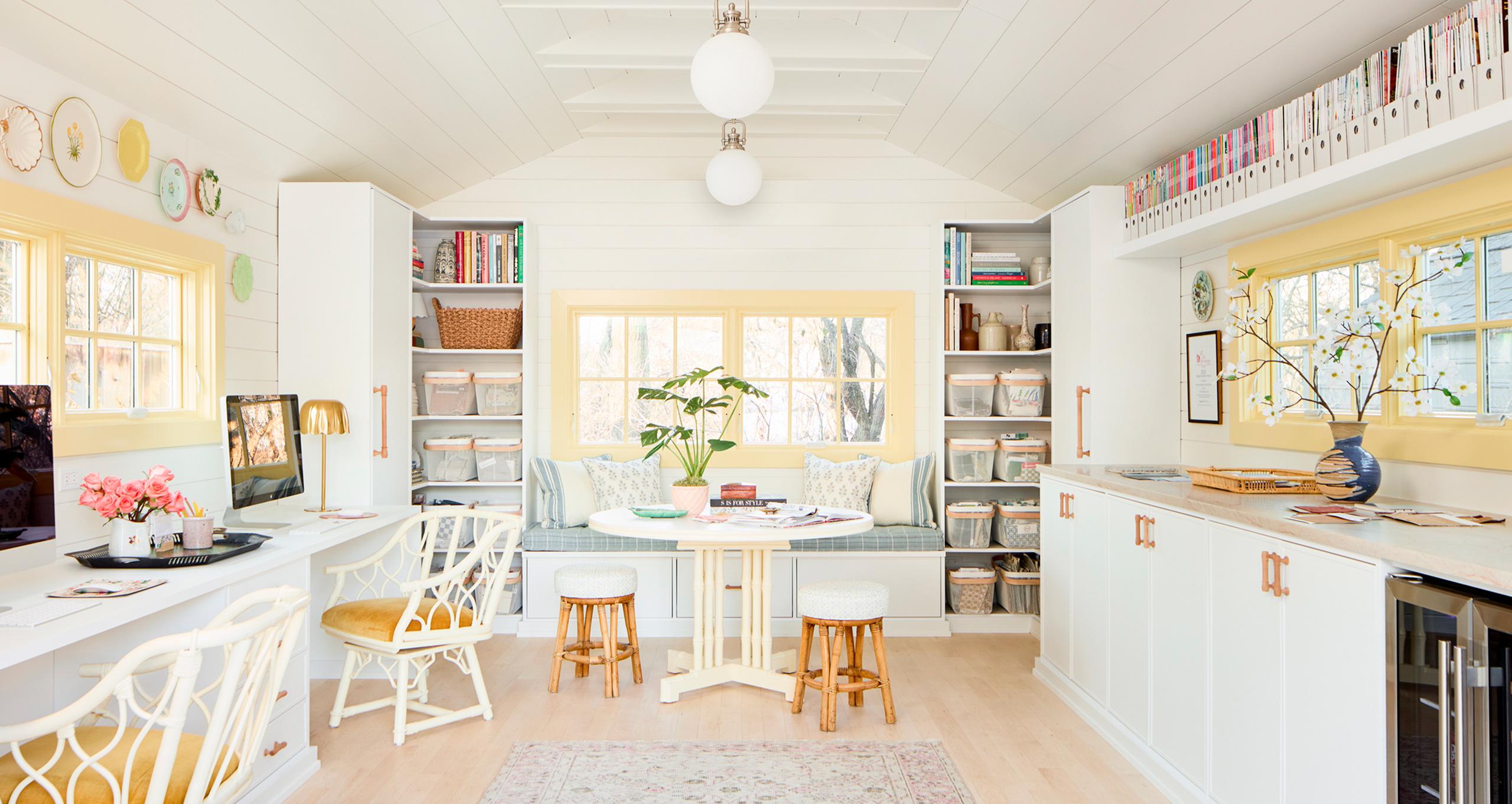 A bright detached garage converted into a professional design studio by California Closets, featuring custom Dove white cabinetry, dual workstations, a cushioned window bench, and organized open shelving for interior design samples.